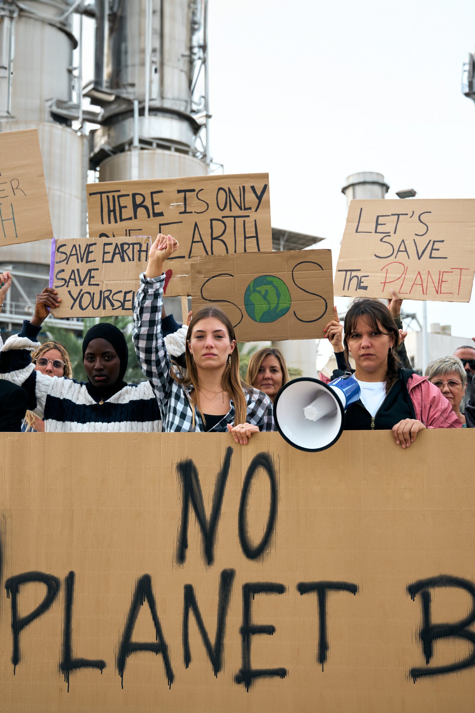 Vertical. Group of people of different ages and nationalities with arms raised in protest against pollution. Pro-earth demonstration with climate change banners looking at camera with serious face